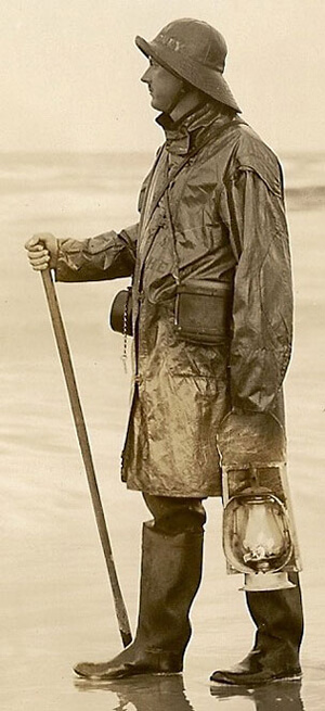 An old photo of a man on a beach in rain gear holding a lantern and walking stick
