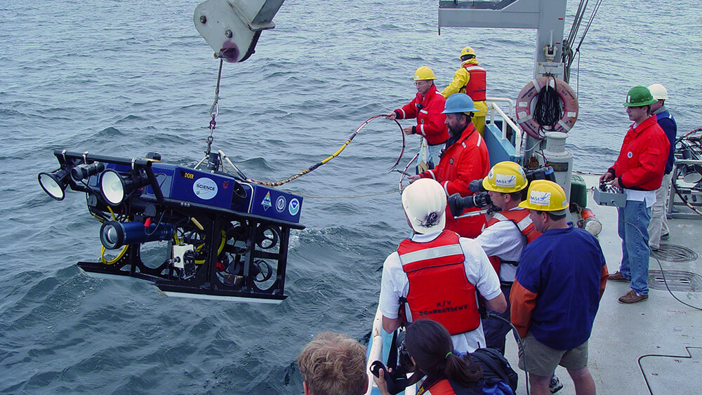 People on the deck of a ship work to lower an rov into the water