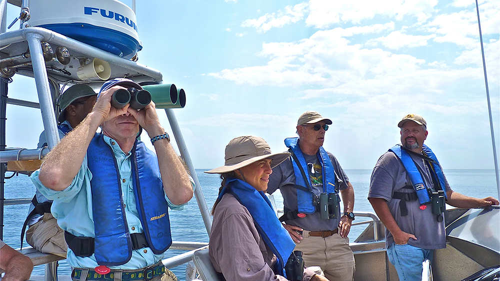 a person looking through binoculars on a research vessel with other people in the background